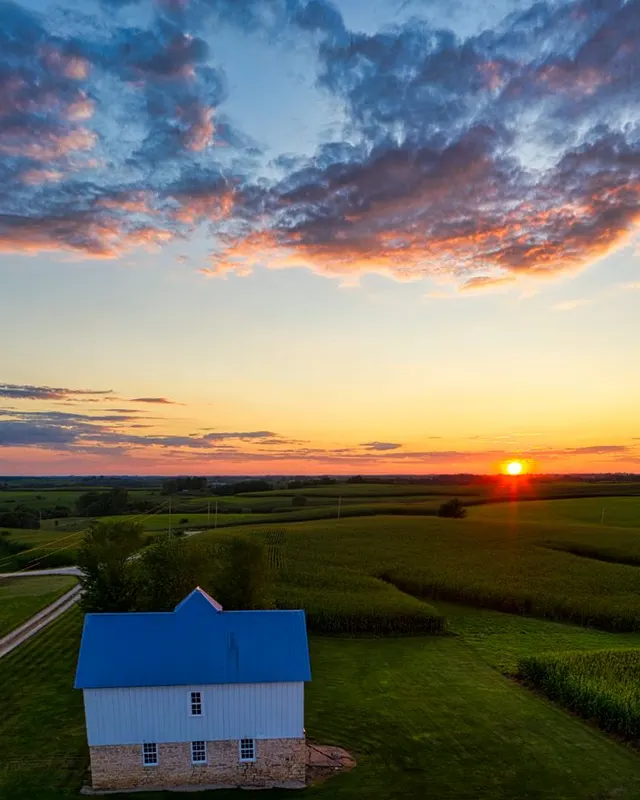 Aerial view of a tidy white farmhouse with a blue roof beside green cornfields at sunset